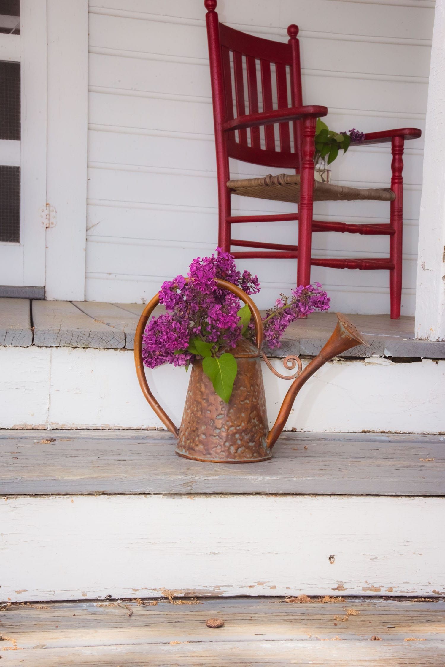 A watering can filled with purple flowers sits on a porch next to a rocking chair