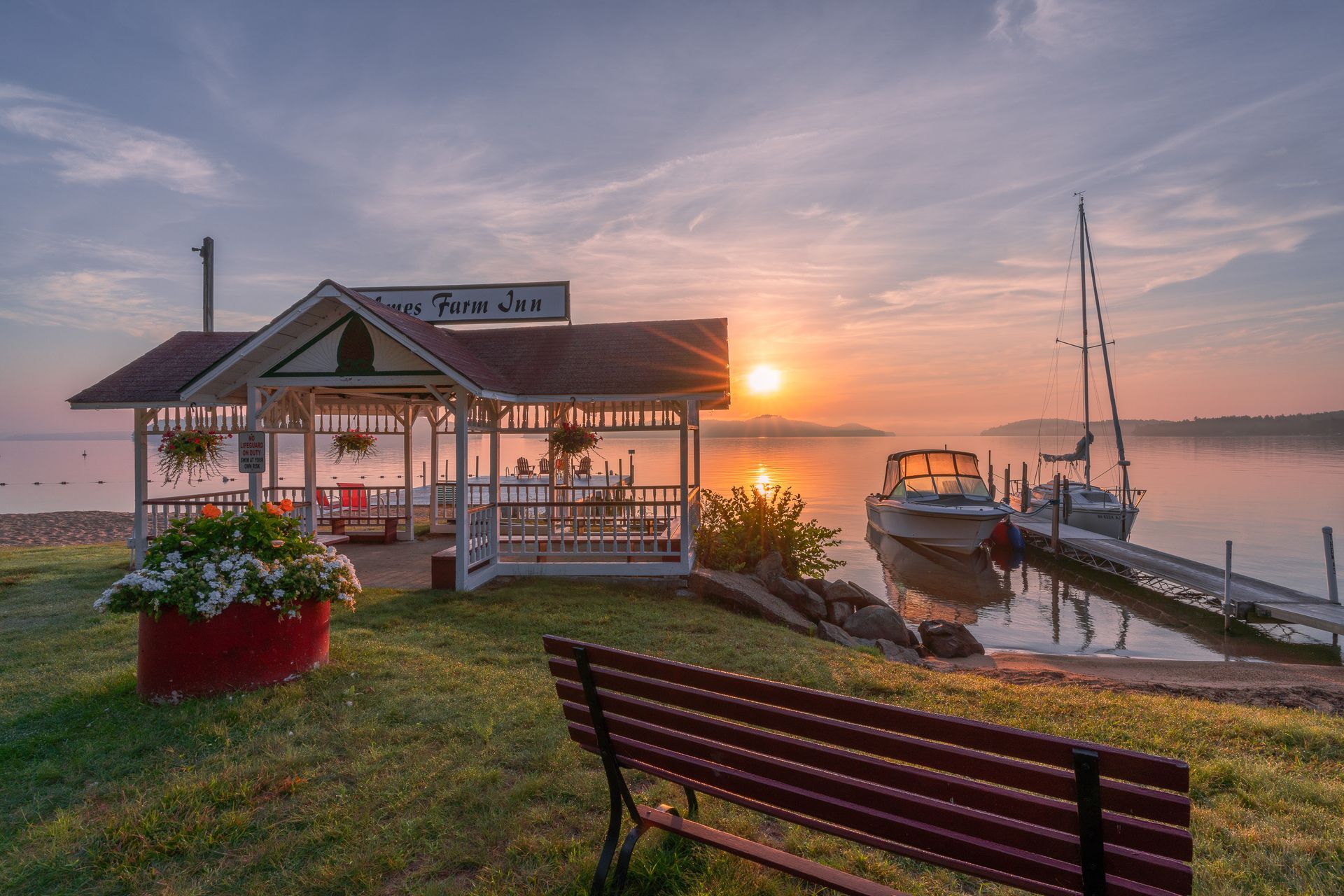 Sunrise over a calm lake. A gazebo, boats, and a bench are in view. Orange and pink hues reflect on the water.