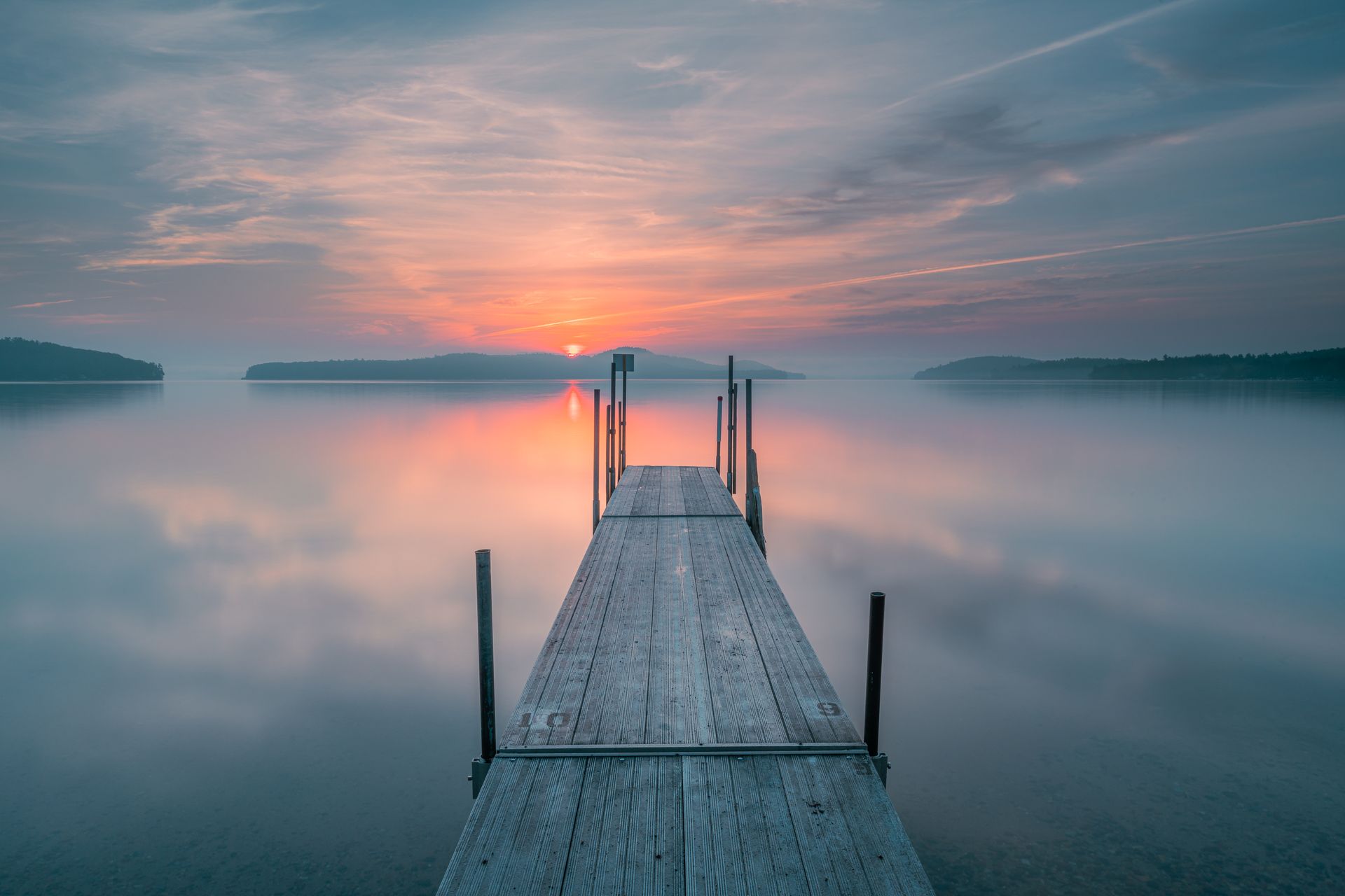 Wooden pier extending into a still lake at sunrise; the sun's reflection and colors of orange and pink on the water.