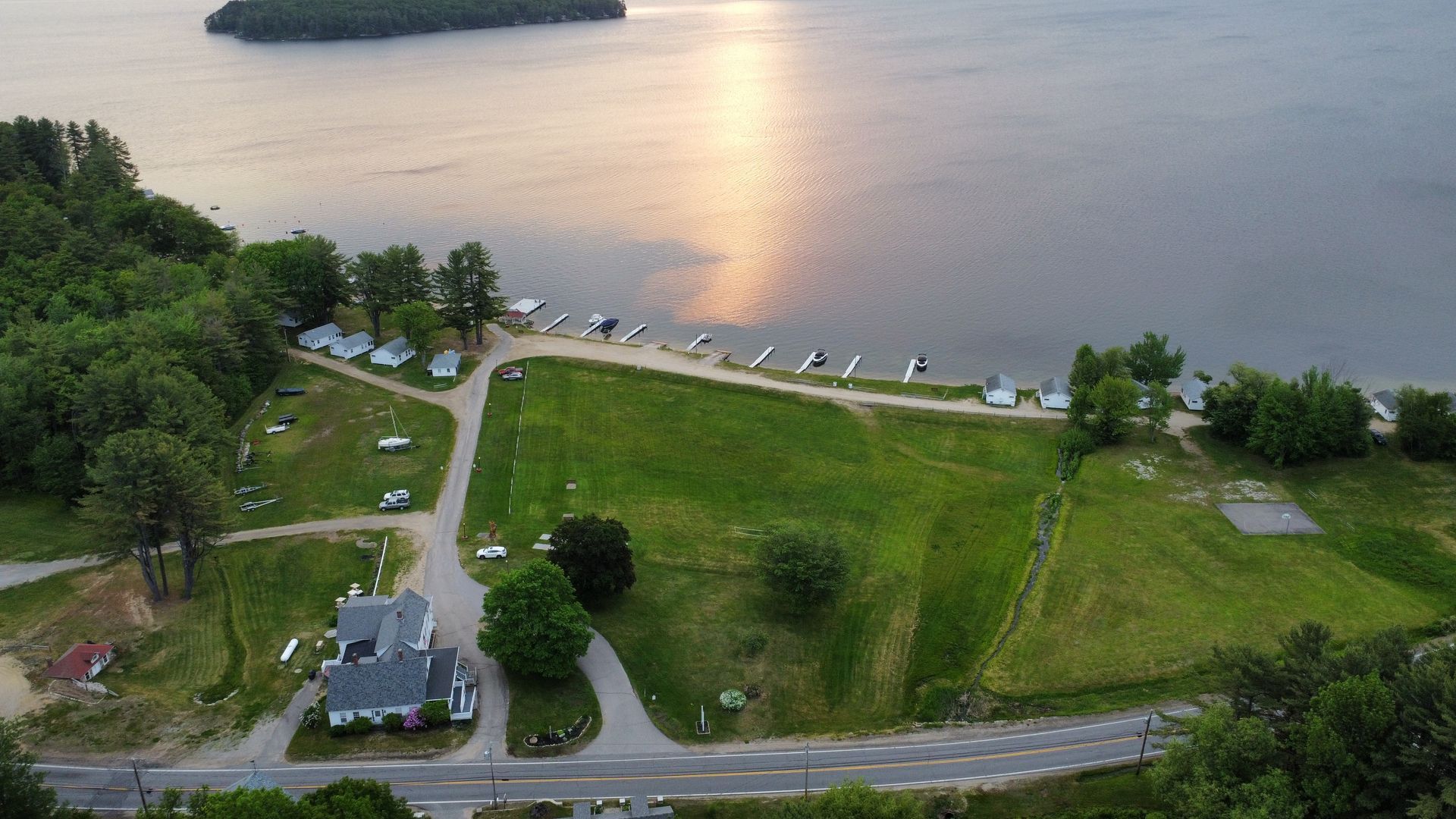Aerial view of a lakefront property at sunset. A grass field leads to a shoreline dotted with boats and small cabins.