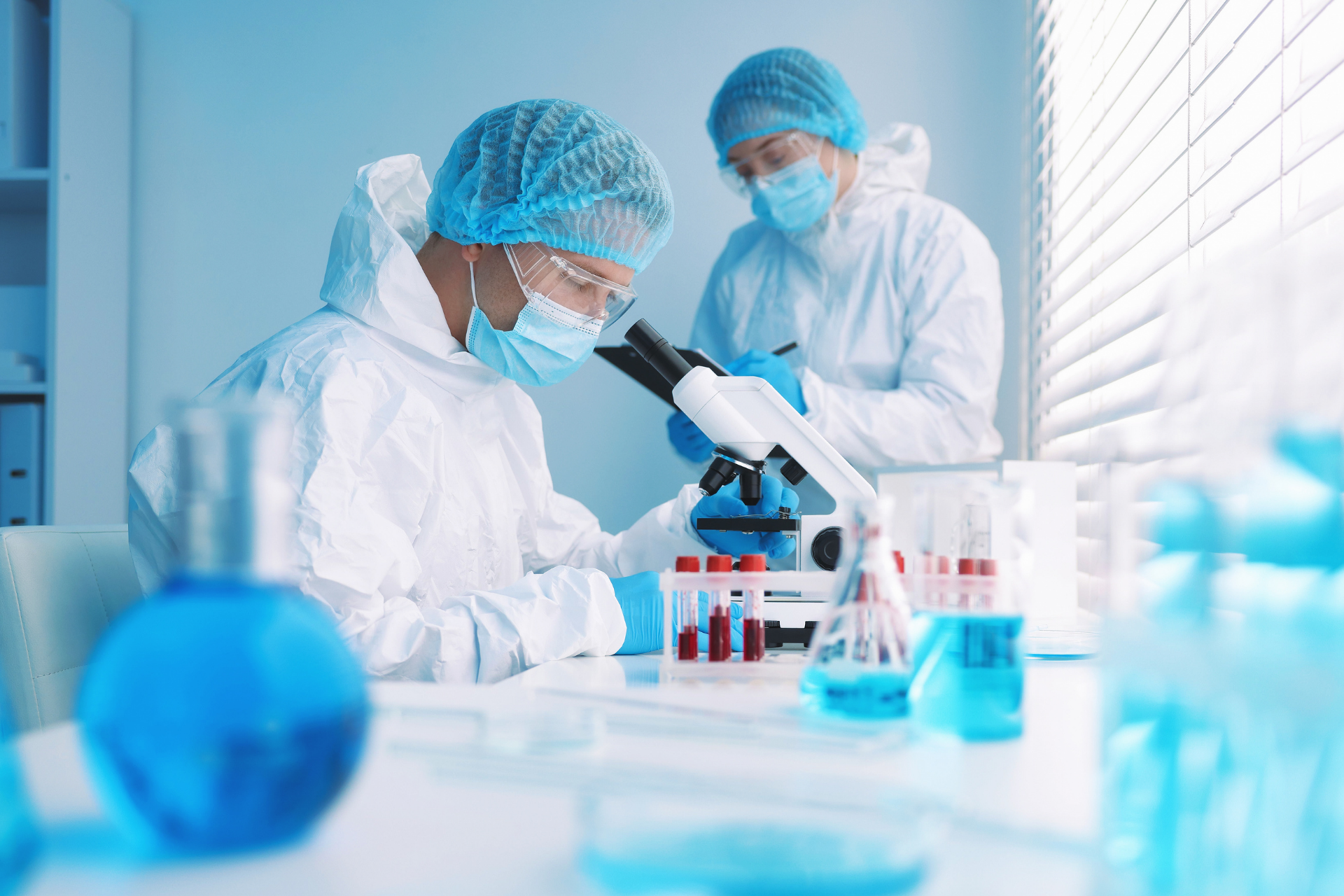 Two lab workers in protective suits examine samples with a microscope in a blue-lit lab.