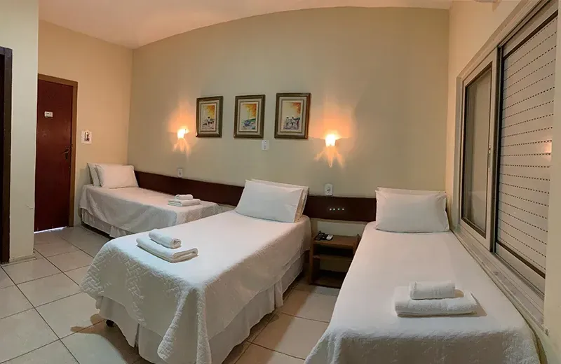 Quarto de hotel com três camas, lençóis brancos, paredes em tons neutros, obras de arte emolduradas e uma pequena mesa de cabeceira.