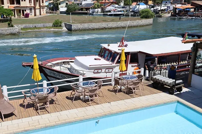 Um barco atracado perto de um deck com mesas, cadeiras e uma piscina. Água azul e prédios ao fundo.