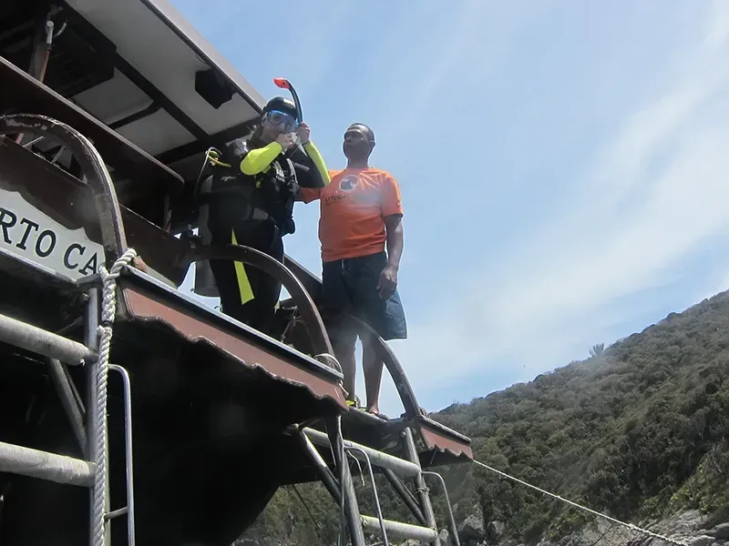 Duas pessoas em um barco perto de uma costa rochosa; uma com equipamento de mergulho, a outra com uma camisa laranja.
