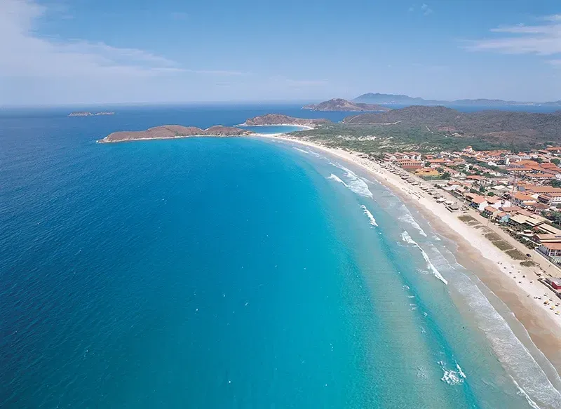 Vista aérea do oceano turquesa encontrando uma praia de areia, edifícios e uma costa montanhosa.