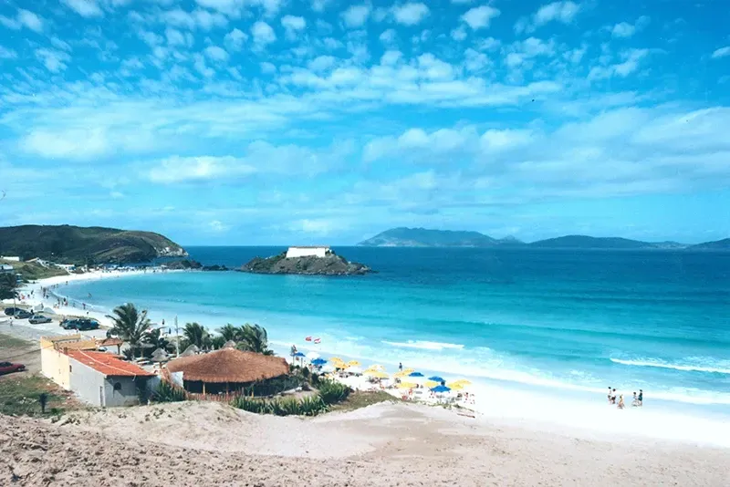 Praia com areia branca, água turquesa, céu azul e uma pequena ilha com uma estrutura.