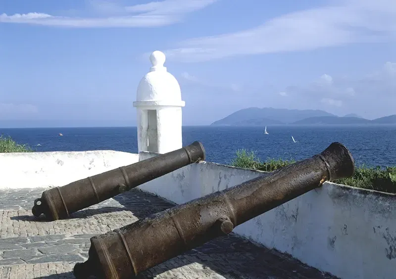 Dois canhões em uma parede branca com vista para o oceano azul e ilhas distantes sob um céu claro.