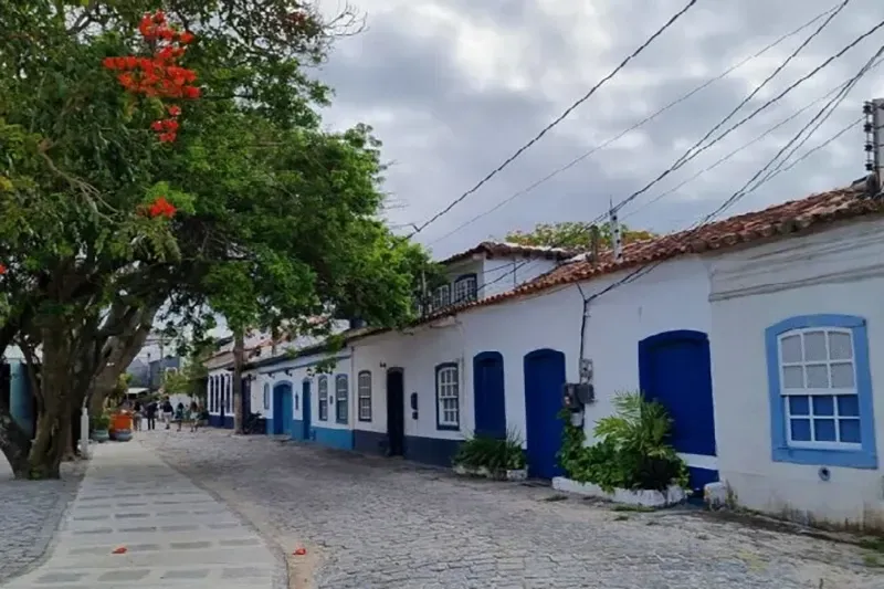 Rua de paralelepípedos ladeada por prédios brancos, portas e venezianas azuis, sob um céu nublado.
