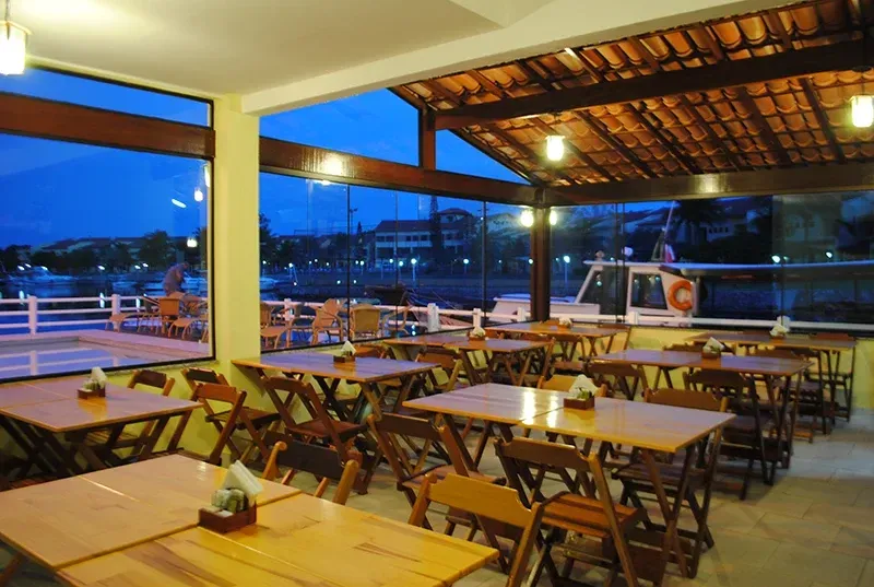 Restaurante com mesas de madeira, paredes de vidro e teto de telhas com vista para a orla ao entardecer.