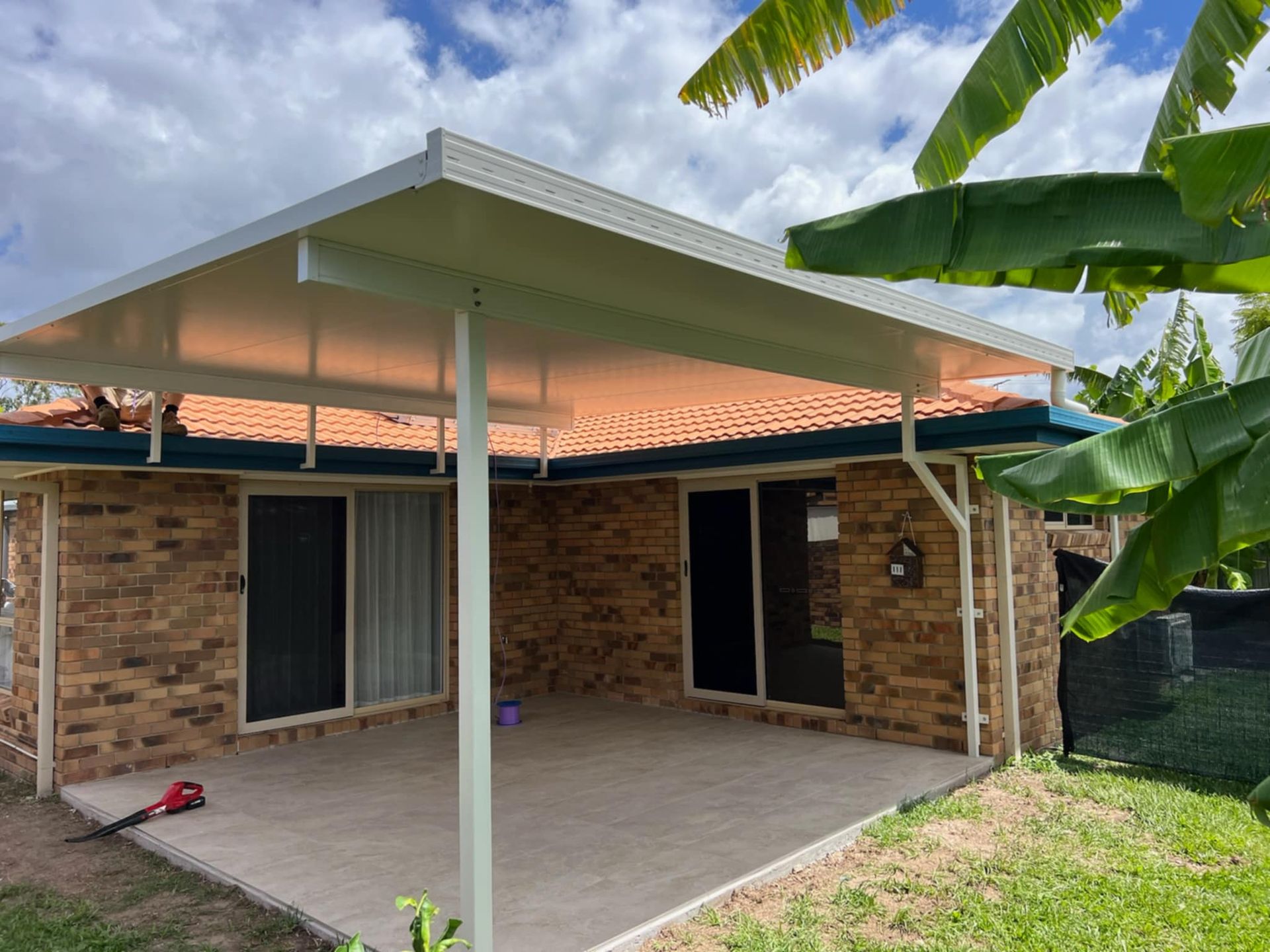 Patio with brick walls and a white roof, under a blue sky, with greenery on the right.
