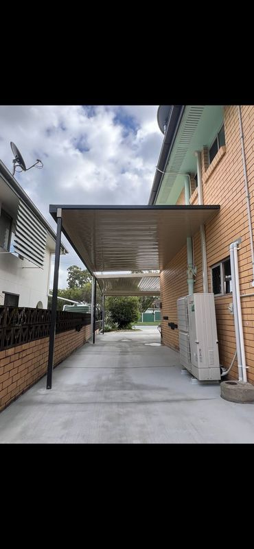 A covered carport with a concrete driveway between a brick wall and a building. Blue sky above.