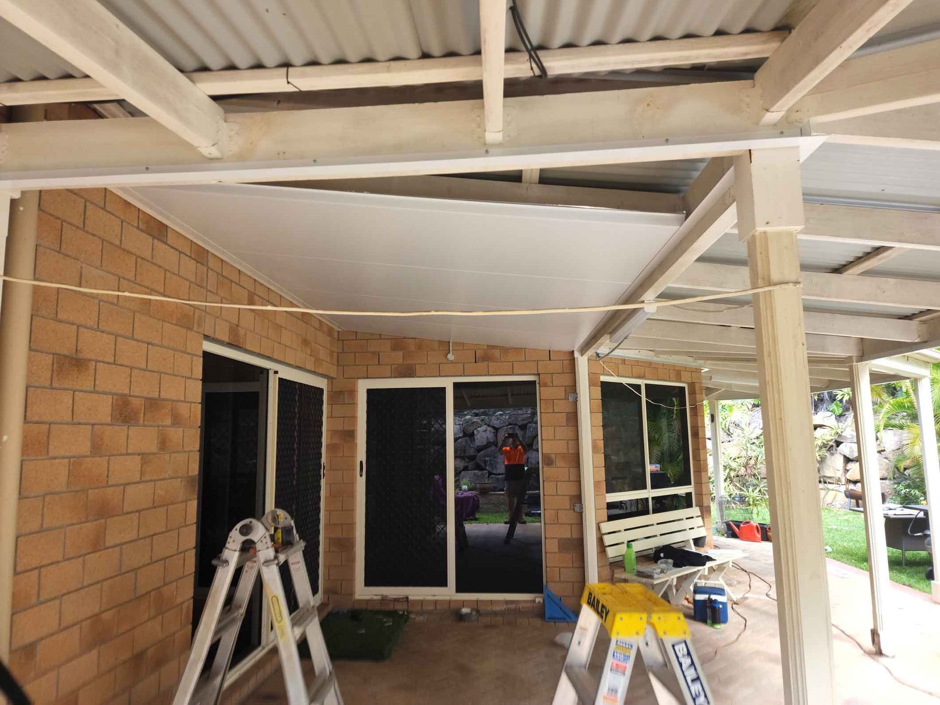 A brick house with a covered patio. White ceiling, sliding glass doors, a person visible inside, and ladders present.