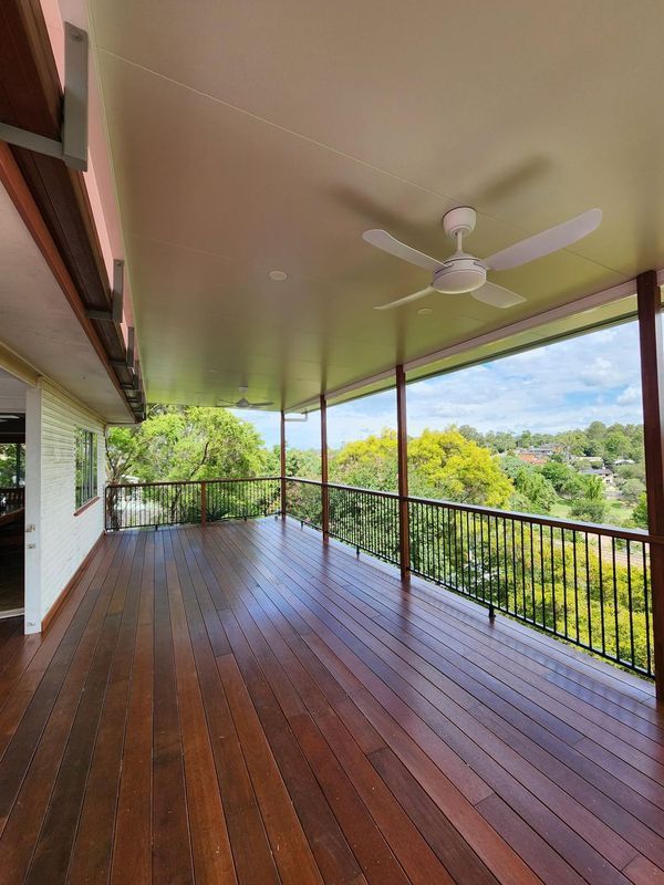 Wooden deck with black railing and white ceiling fan, overlooking trees and a green landscape.