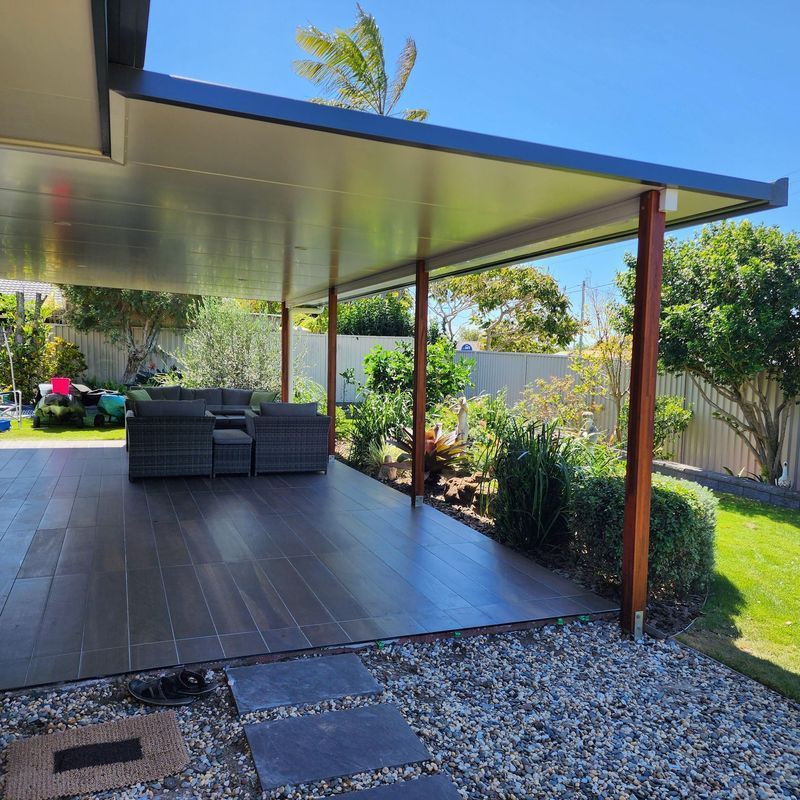 Patio with gray tile flooring and a covered roof, supported by wooden posts. Outdoor seating and plants are visible.