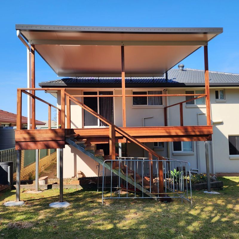 Wooden deck with stairs, built-in patio cover, and railing. Attached to the back of a two-story beige house.