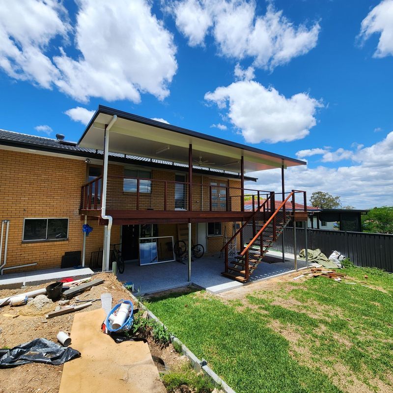 Two-story house with wooden deck and stairs, concrete patio, green grass, and blue sky with clouds.