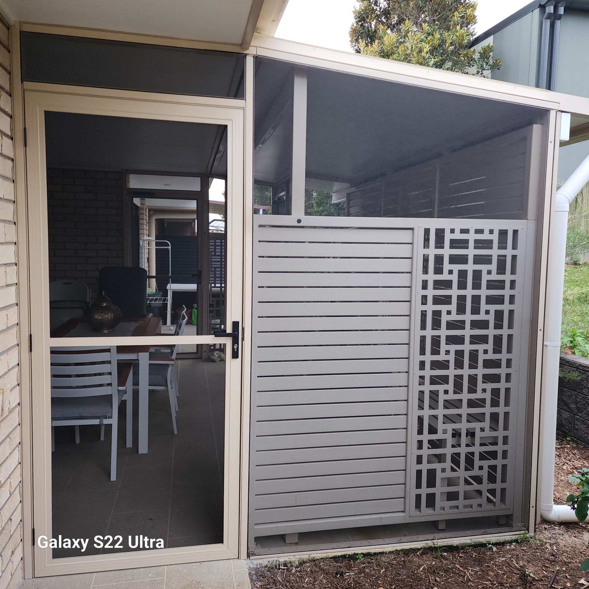 Covered patio with a beige roof and support beams. Black shade screens and beige decorative gate are in view.