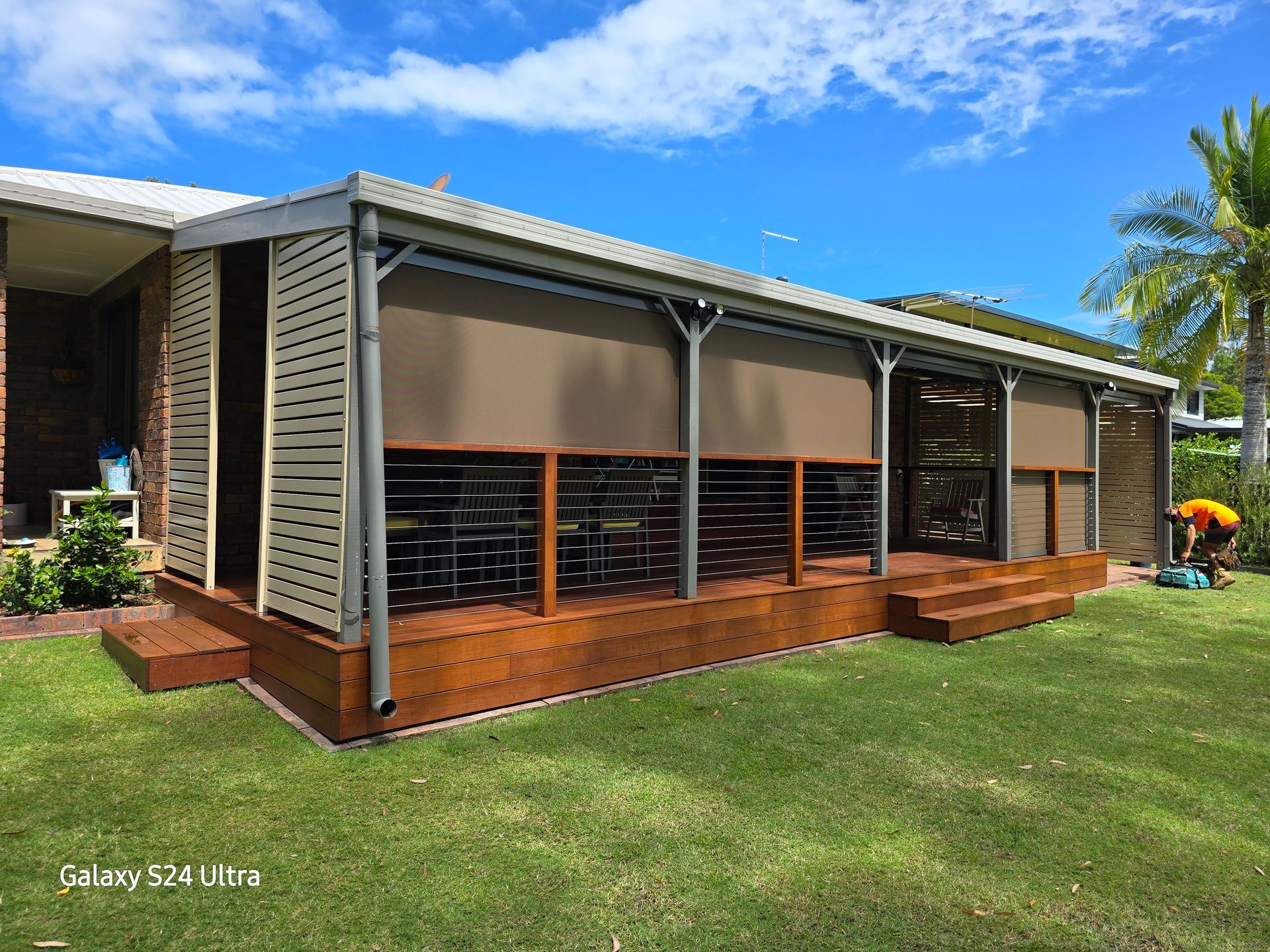 Outdoor deck with brown sun shades, wooden slats, and a grassy yard under a blue sky.