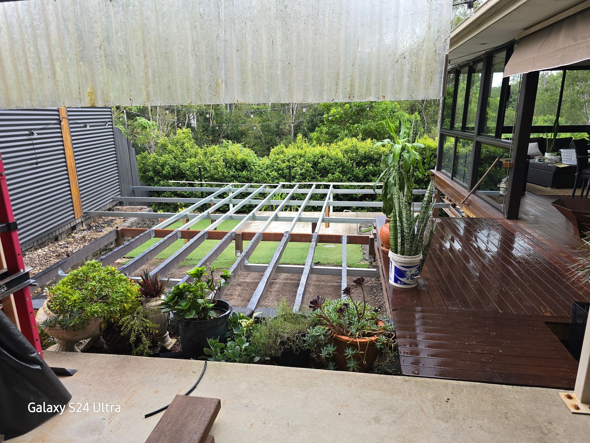 Framed deck under construction, surrounded by greenery and a corrugated metal fence, with a translucent roof overhead.