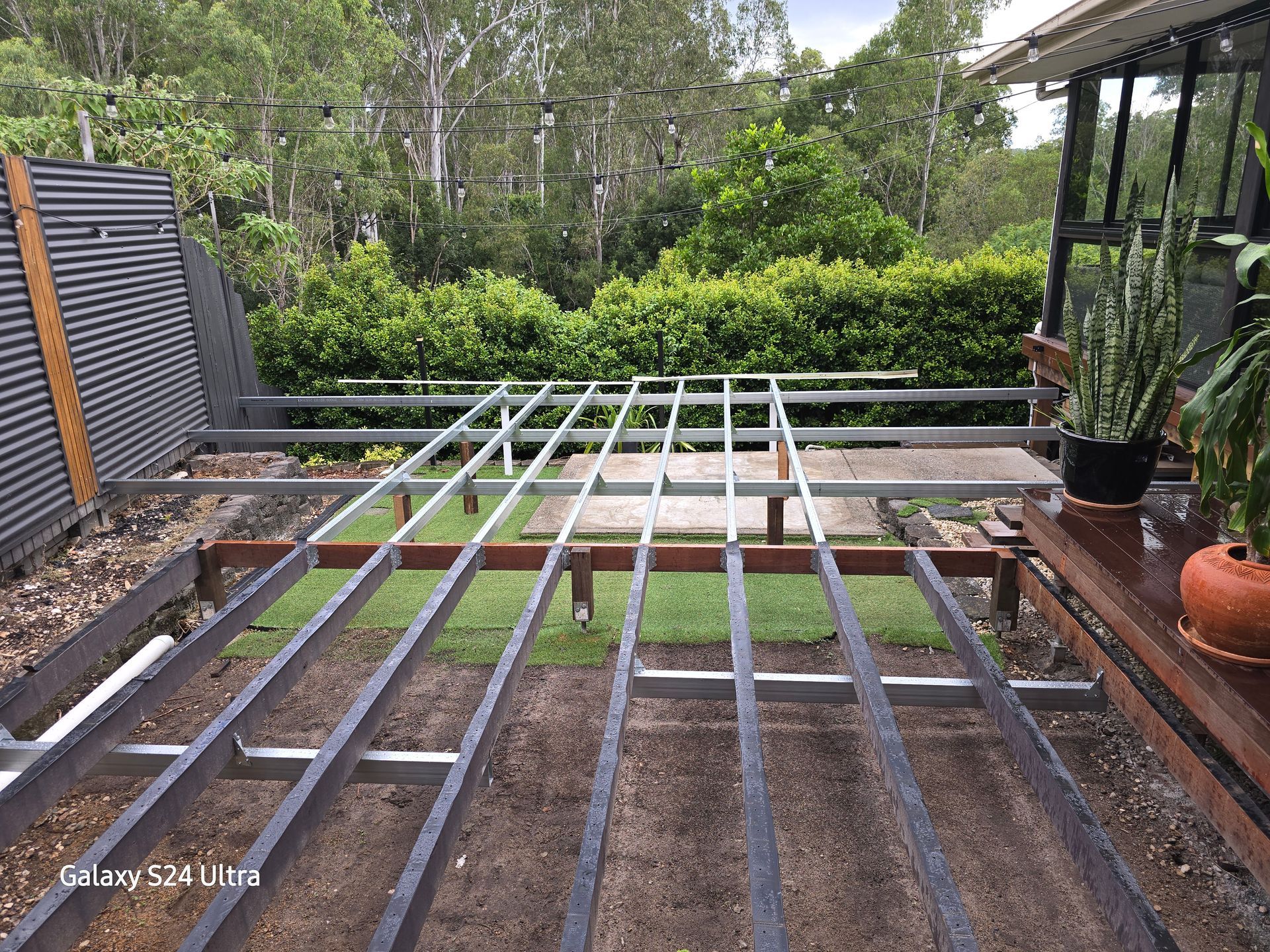 Metal deck frame under construction outdoors, surrounded by foliage and existing deck.