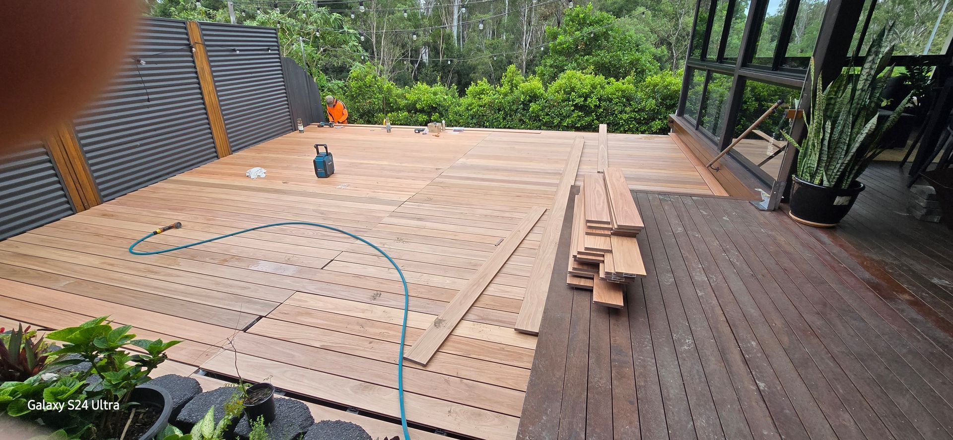 Deck under construction with scattered wooden planks. Green hose, potted plants and trees in background.