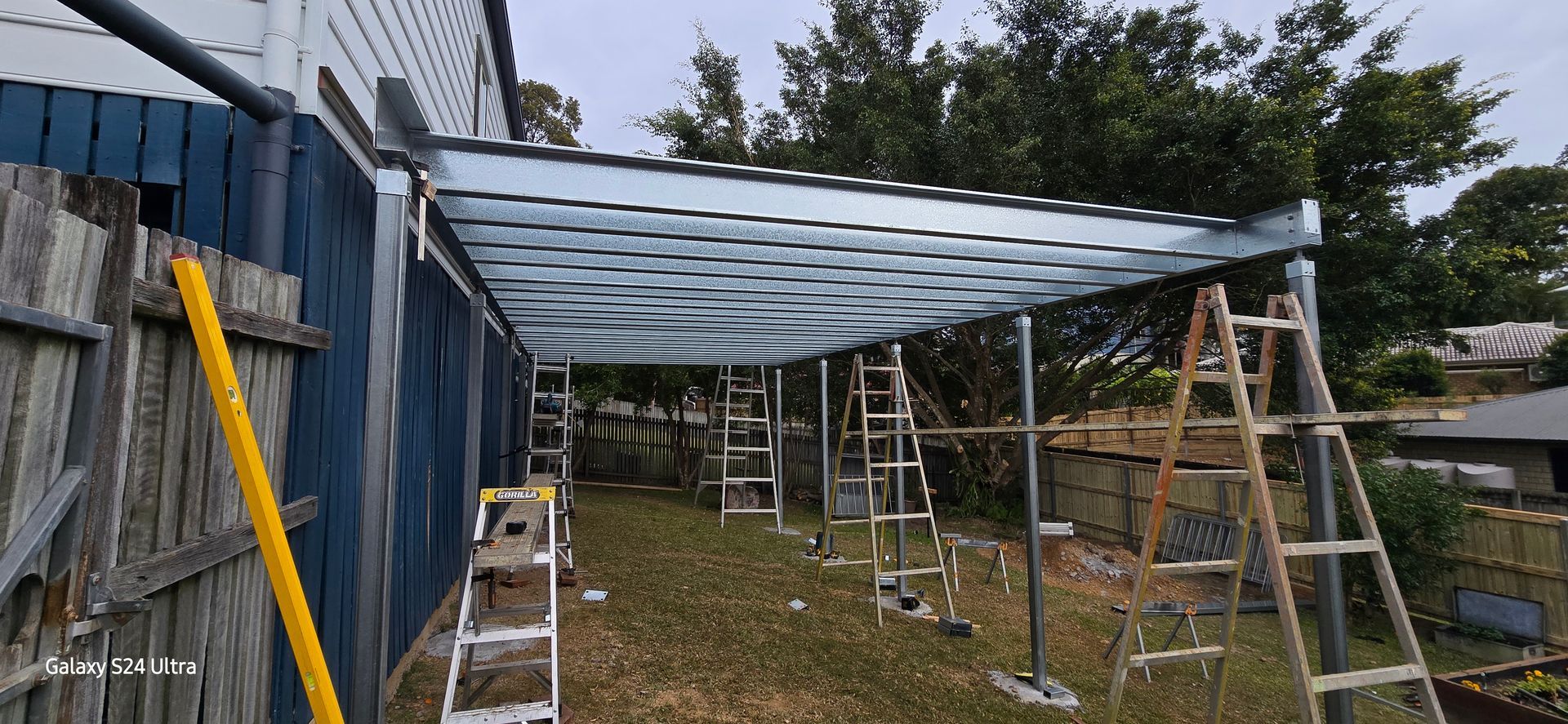 Construction of a metal awning attached to a house with ladders and scaffolding on grass.