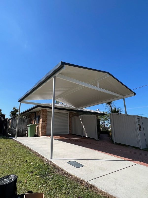 Carport over a driveway in front of a brick house with a green trash can and a clear blue sky.