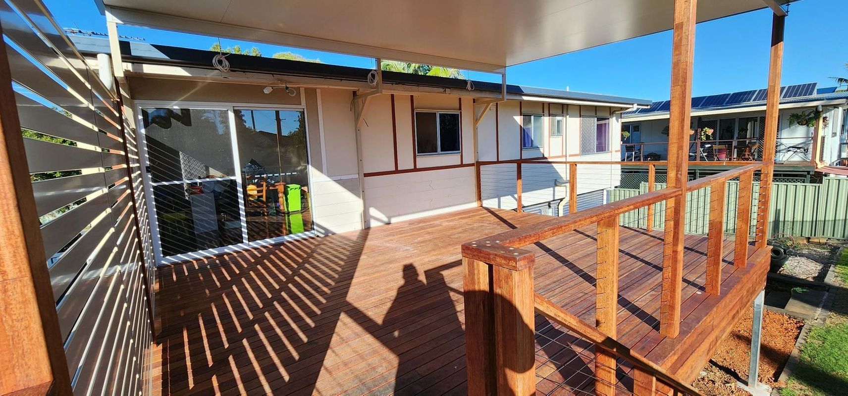 Wooden deck with railing, shadows, and a view of a house. Blue sky overhead.