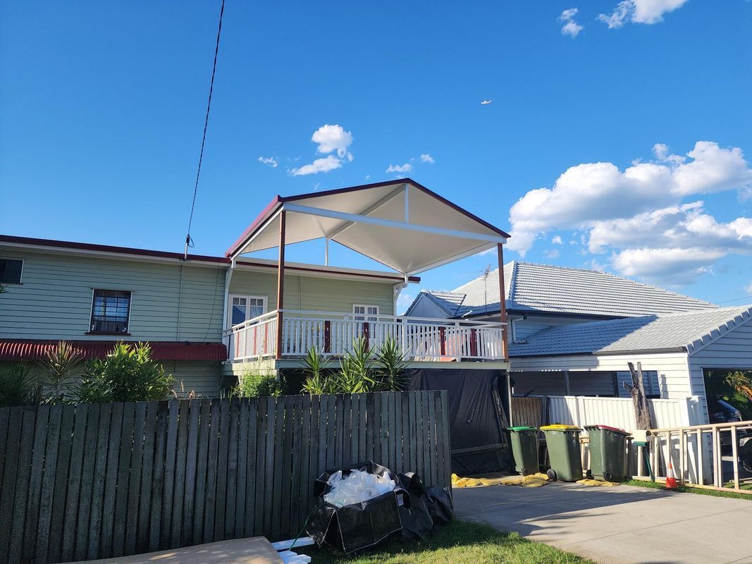 Green house with white deck, tan awning, blue sky, wooden fence. Garbage cans.