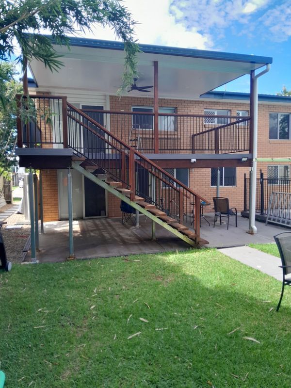 Two-story brick building with deck and stairs. Brown railing, grass yard, and a light blue sky.