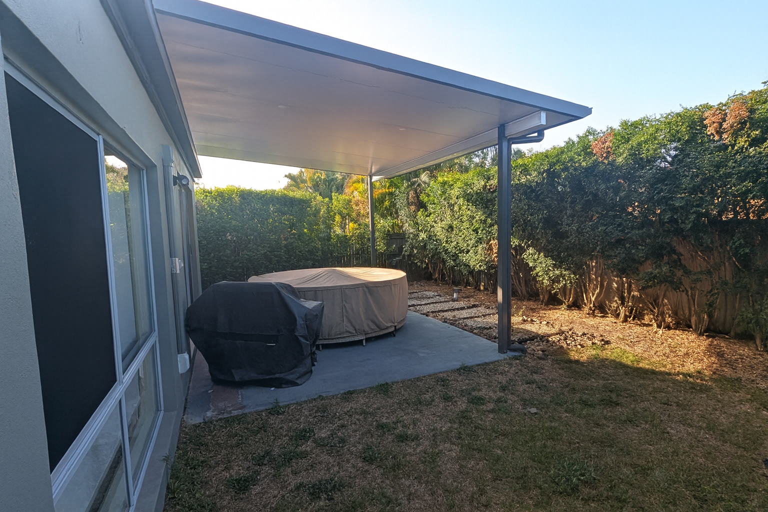 Patio with covered hot tub, concrete, gray roof overhang, and building in the background.