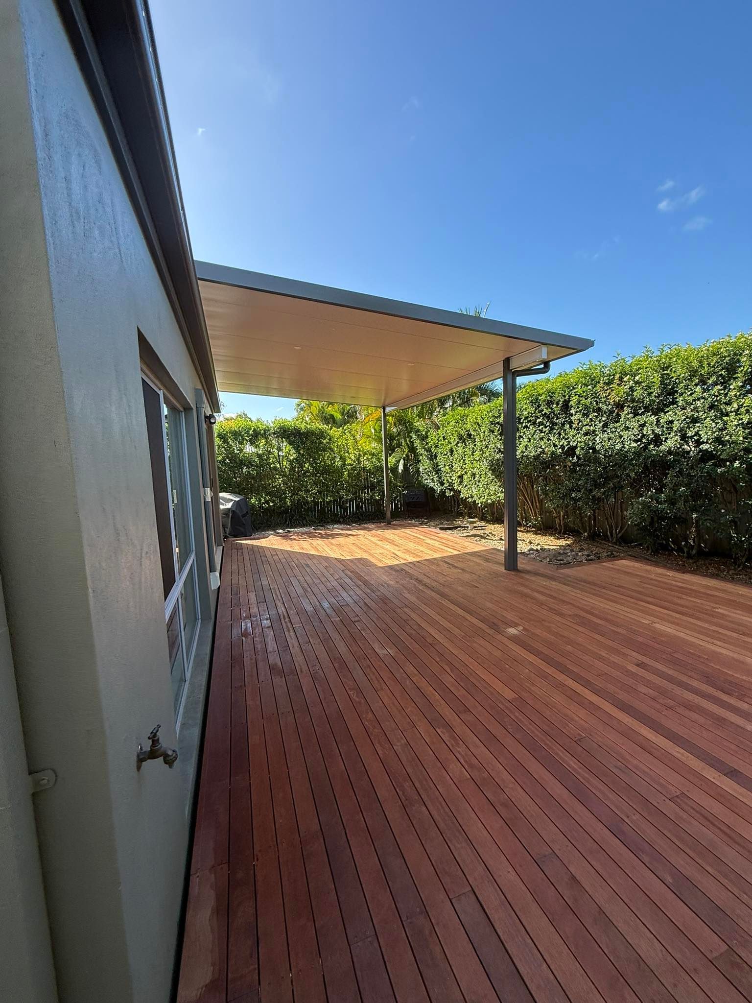 Wooden deck with a covered area, surrounded by lush green foliage and a beige building.