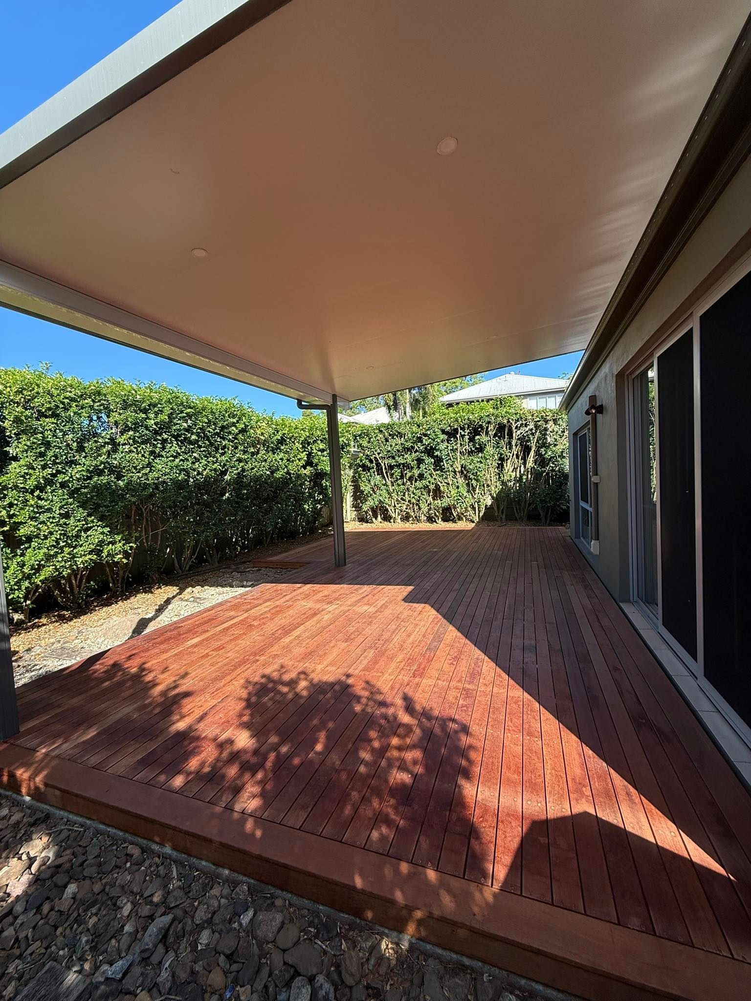 Wooden deck with awning, bordering a hedge and house with sliding doors.