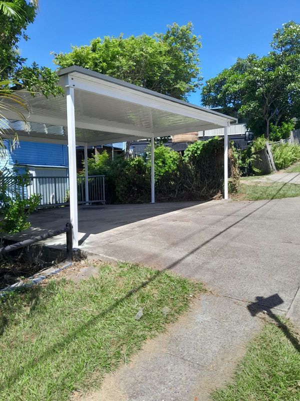 Carport with white supports, gray roof, on a driveway with a curb and grass. Sunny day.