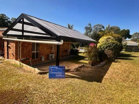 A brick house with a carport under a blue sky. A sign is placed in the grass.