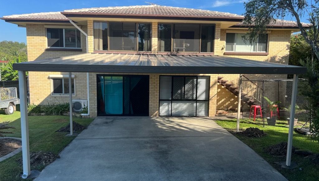 Two-story house with carport. Beige brick exterior, large windows, concrete driveway, and grassy yard.