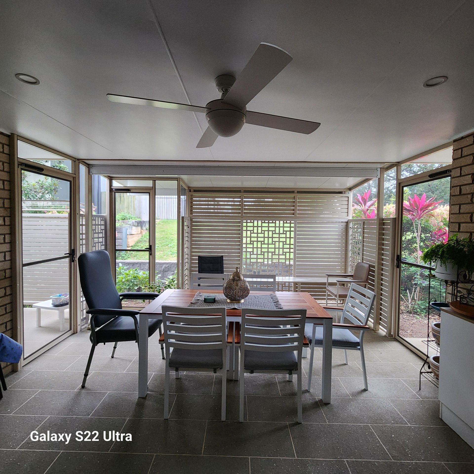 Indoor dining area with table and chairs, enclosed by glass walls and brick, with a ceiling fan.