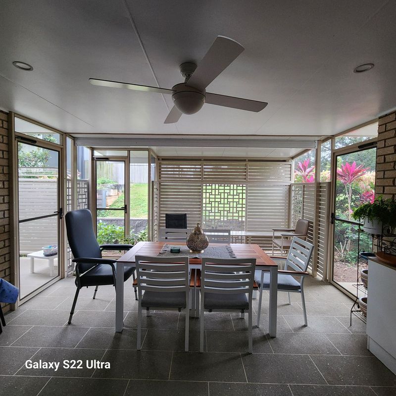 Enclosed sunroom with dining table, chairs, brick accents, and a ceiling fan; windows overlook greenery.