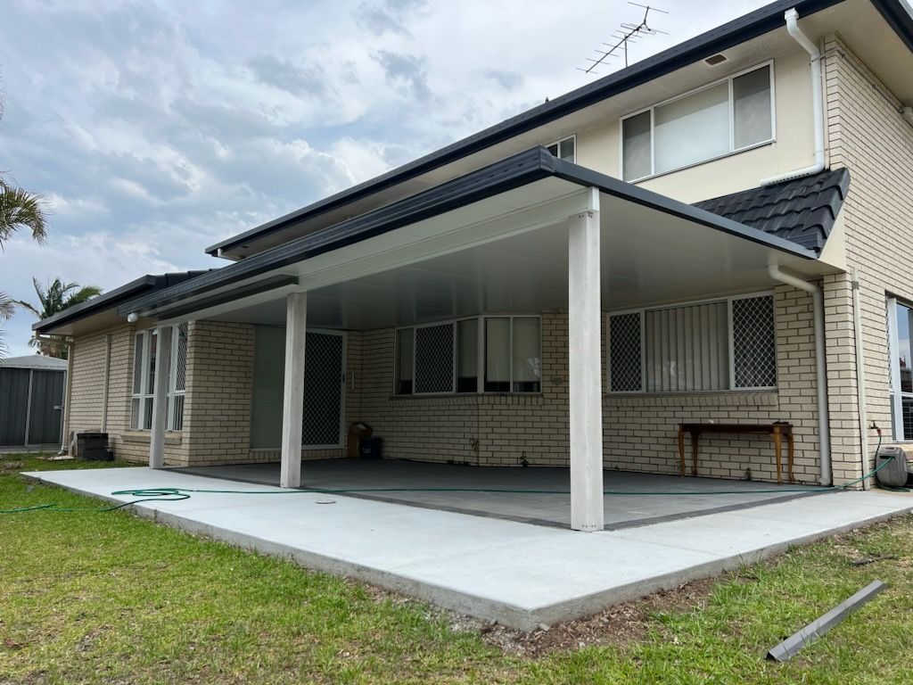 A covered patio with white pillars extending from a two-story brick house, concrete floor, green grass.