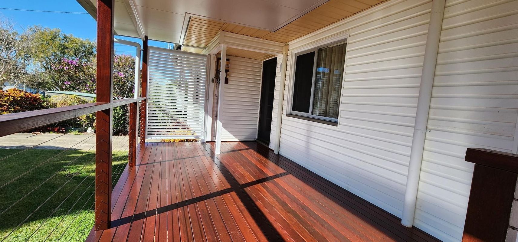 Wooden porch of a house with white siding and a slatted wall, sunny outdoors.