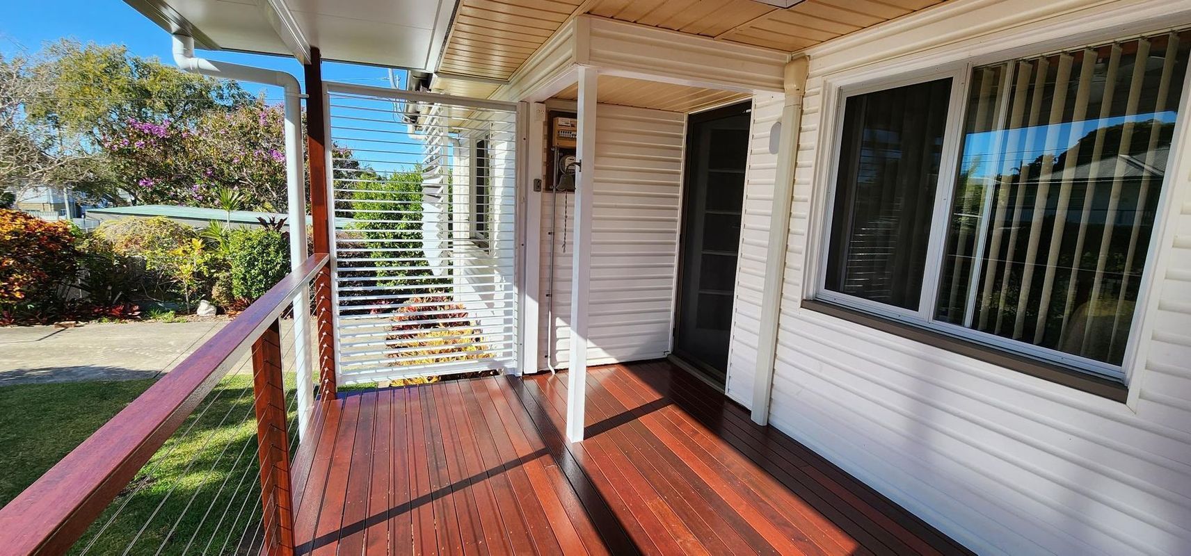 Wooden porch with railing and a white house, lush greenery in the background.