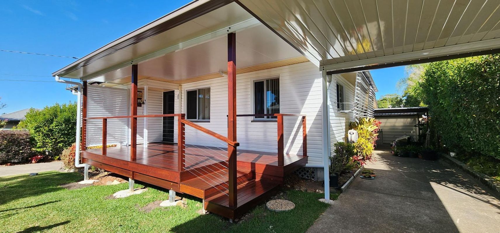 A white house with a brown wooden deck and carport. Green grass surrounds it under a blue sky.