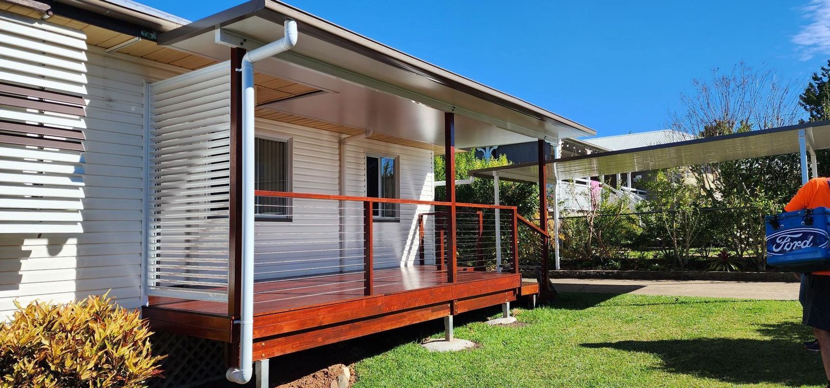 A deck with a red-brown wooden floor and railings extends from a white building. A person stands on the right.