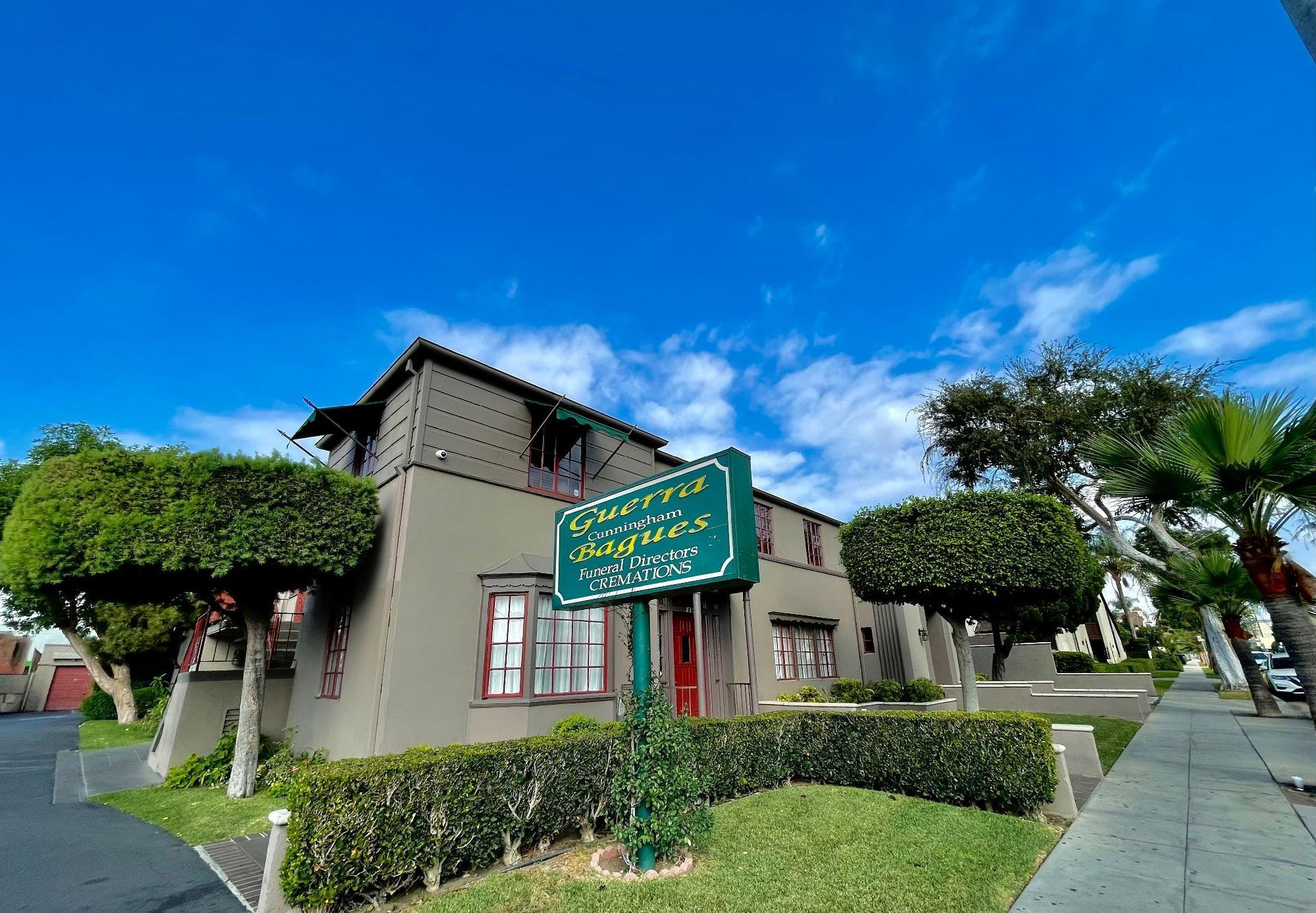 A multi-story beige building with a dark roof and a green sign, surrounded by manicured shrubs under a blue sky.
