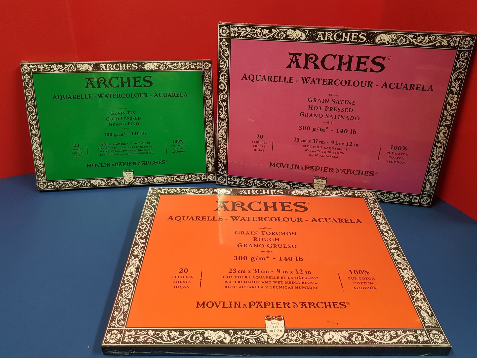 Tre blocchi di carta per acquerelli Arches con copertina verde, rosa e arancione su superficie blu.