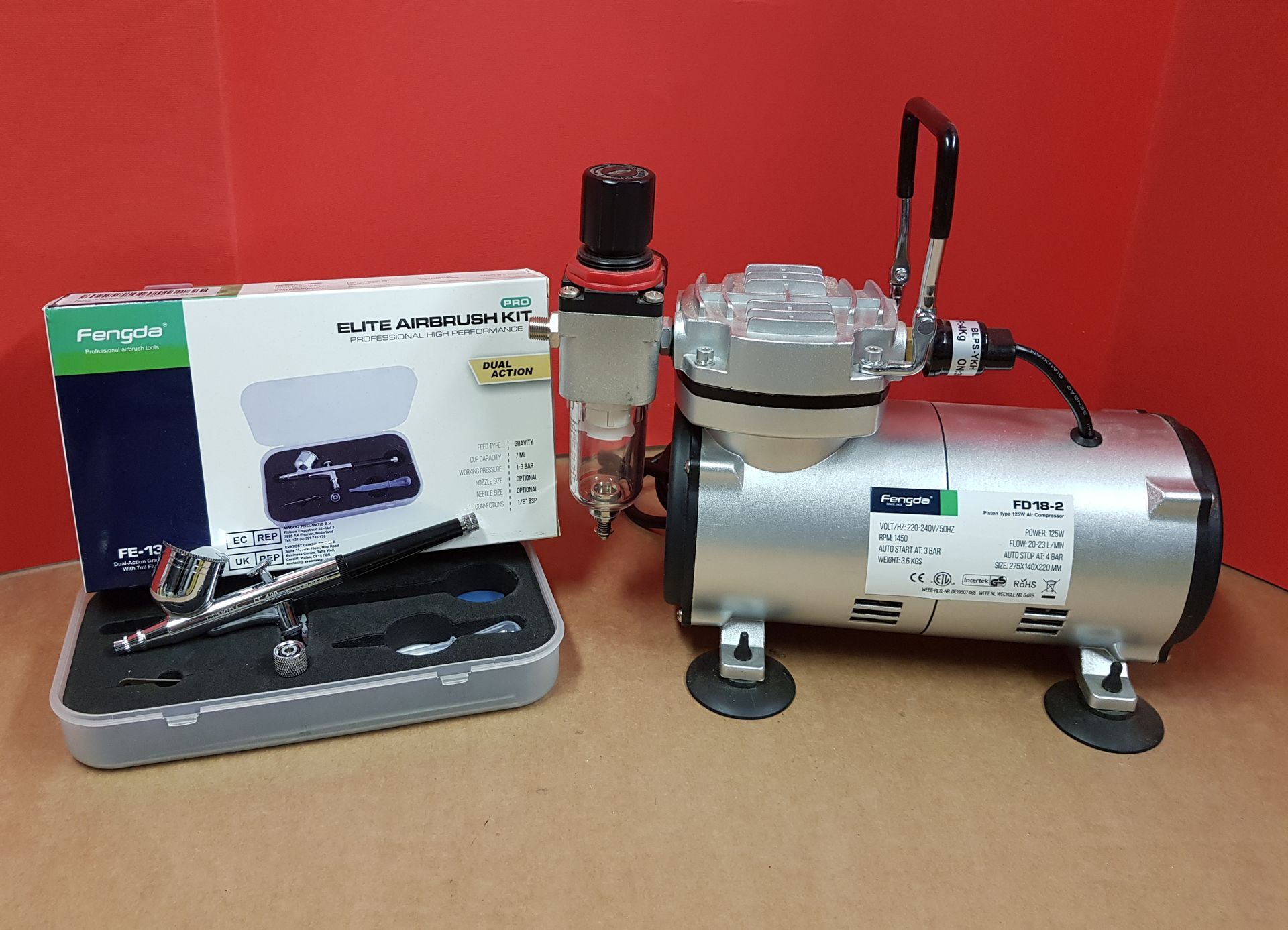 Kit aerografo con compressore, aerografo e accessori su sfondo rosso.