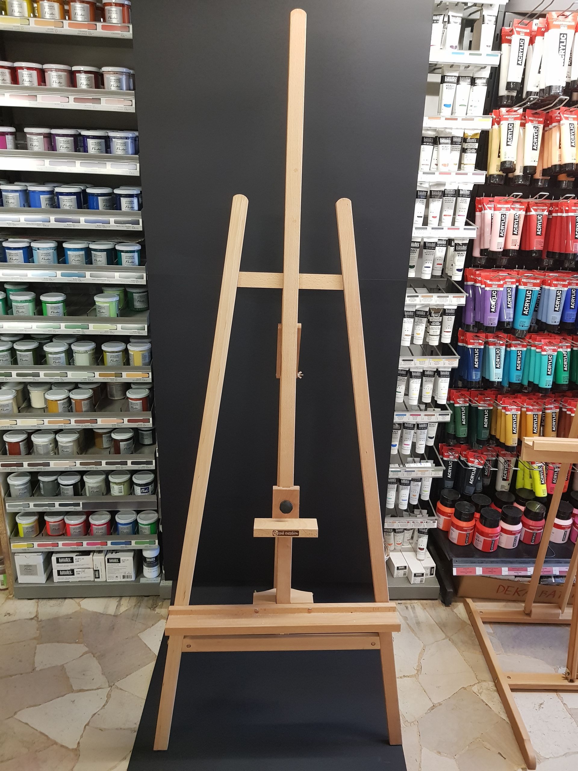Cavalletto in legno per artisti in un negozio di articoli artistici, su uno sfondo nero con scaffali pieni di colori.