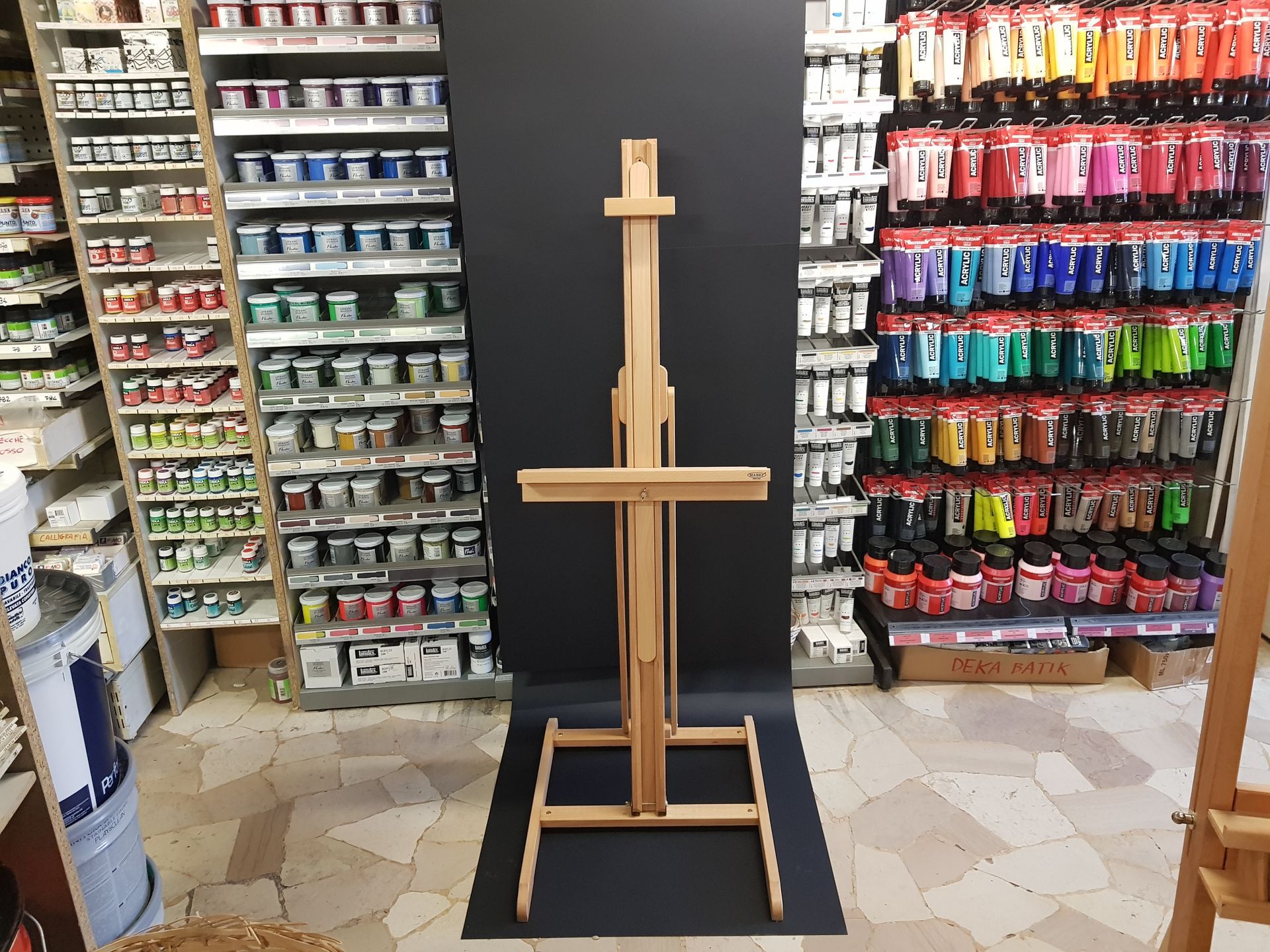 Cavalletto in legno per pittura in un negozio, affiancato da scaffali pieni di colori vivaci.