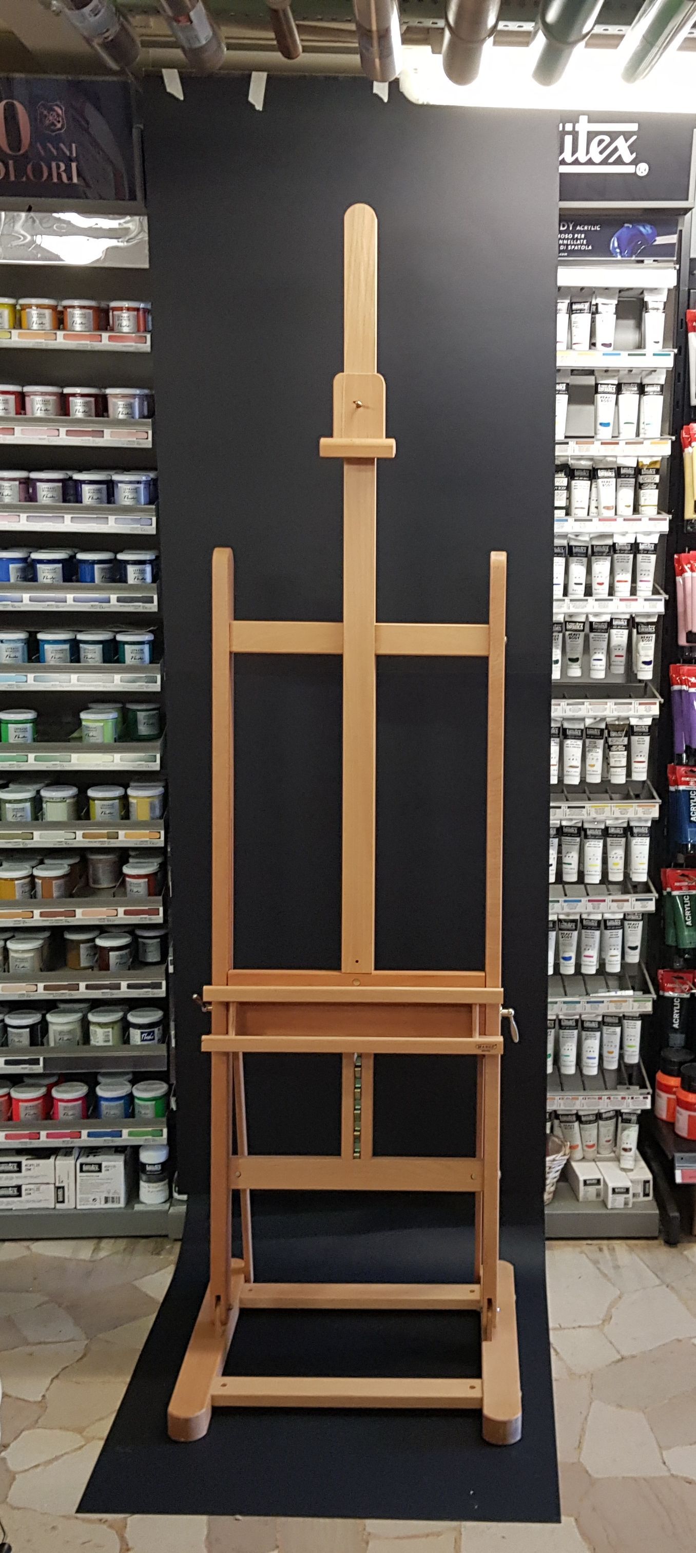Cavalletto da artista in legno esposto in un negozio, affiancato da scaffali pieni di tubetti e barattoli di vernice.