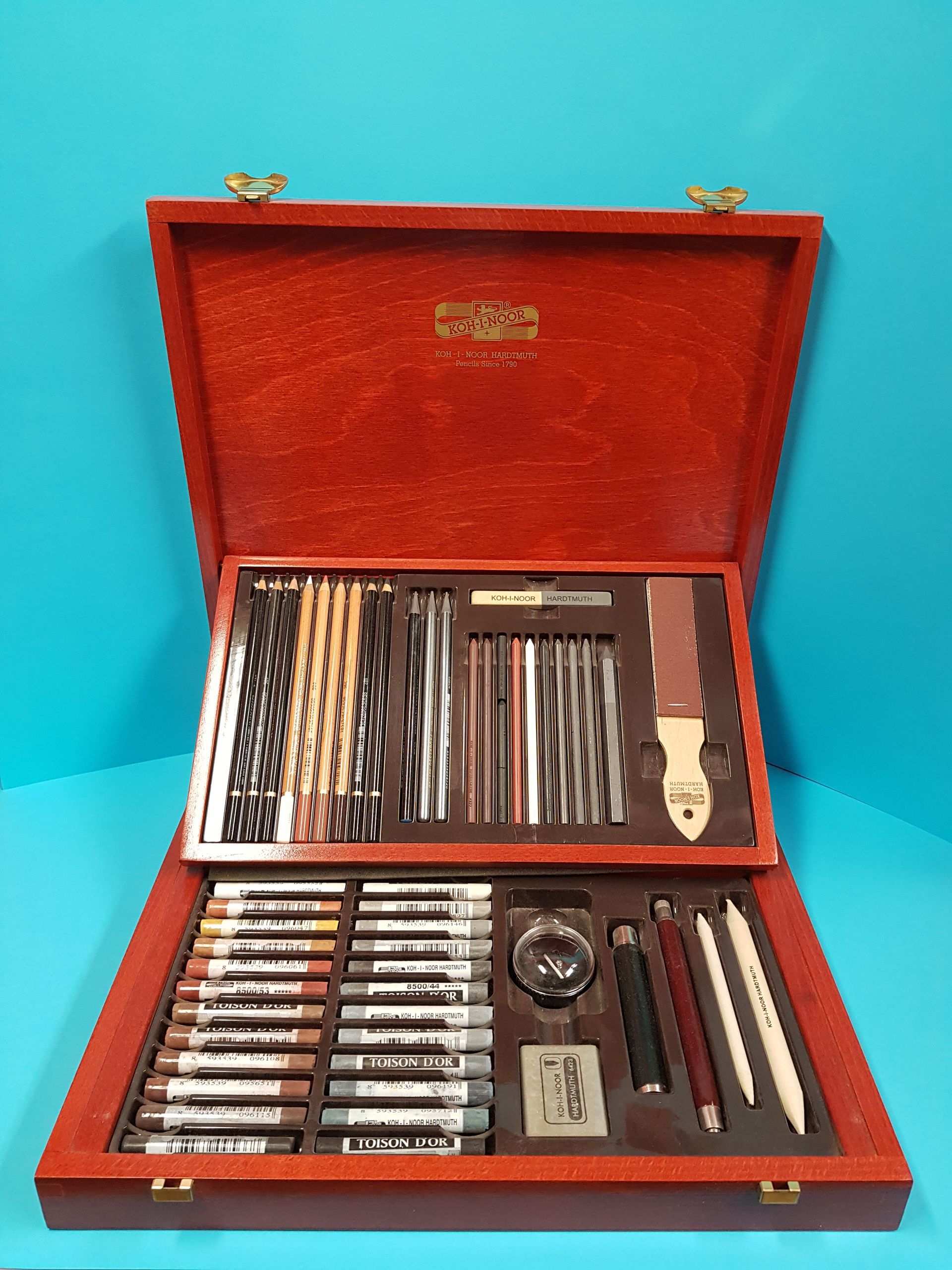 Valigetta in legno per articoli artistici aperta, che rivela matite, carboncini e attrezzi, il tutto su uno sfondo blu.